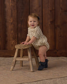 A toddler girl wearing a cream short-sleeve romper with a small floral print sits on a rustic wooden stool in front of a dark wood plank wall.