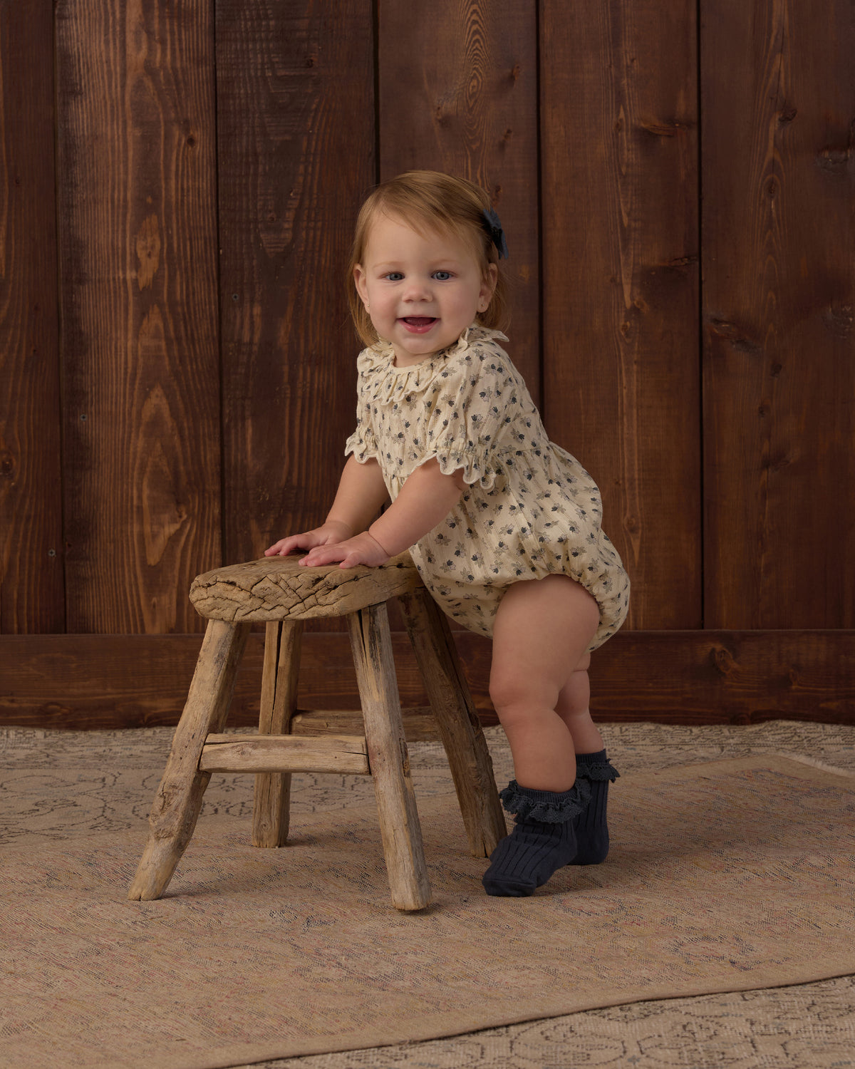 A toddler girl wearing a cream short-sleeve romper with a small floral print sits on a rustic wooden stool in front of a dark wood plank wall.