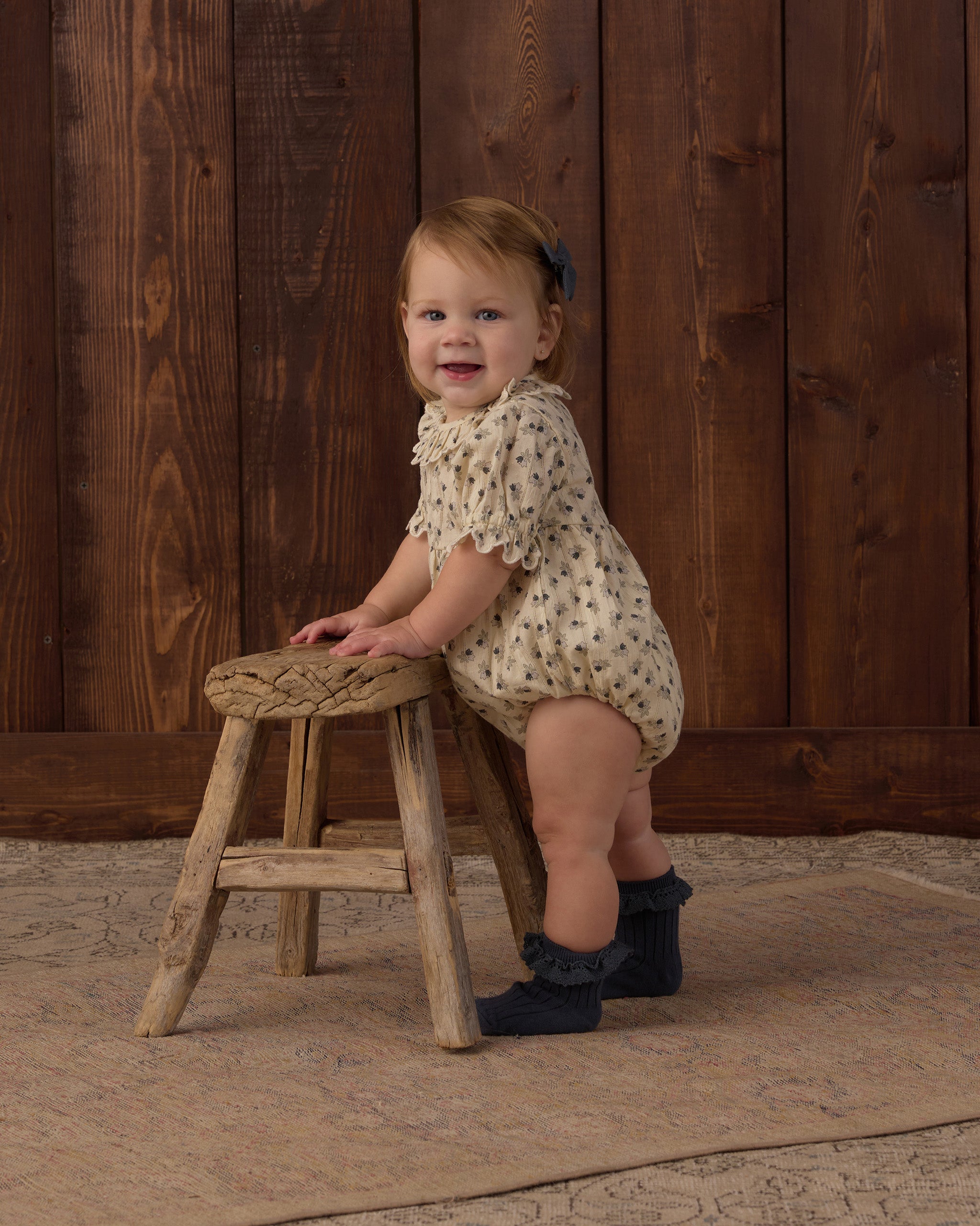 A smiling toddler stands on a rustic wooden stool against a dark wood plank wall, wearing a cream romper with small dark floral print and dark ruffled socks.
