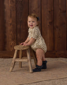 A smiling toddler stands on a rustic wooden stool against a dark wood plank wall, wearing a cream romper with small dark floral print and dark ruffled socks.