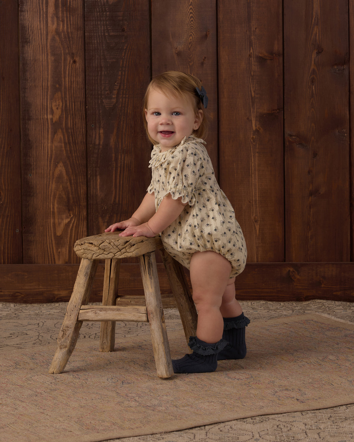 A smiling toddler stands on a rustic wooden stool against a dark wood plank wall, wearing a cream romper with small dark floral print and dark ruffled socks.