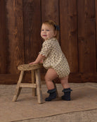 A toddler stands on a rustic wooden stool in front of a dark wood plank wall, wearing a cream short-sleeve romper with a small dark pattern and dark ruffled booties.