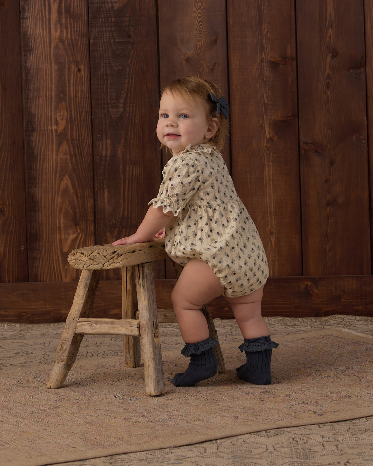 A toddler stands on a rustic wooden stool in front of a dark wood plank wall, wearing a cream short-sleeve romper with a small dark pattern and dark ruffled booties.