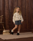 A young girl with curly light brown hair stands in a rustic room with dark wood paneling, wearing a cream knit cardigan with embroidered dark blue star motifs, a dark blue patterned pleated skirt, white ruffled socks, and black shoes.