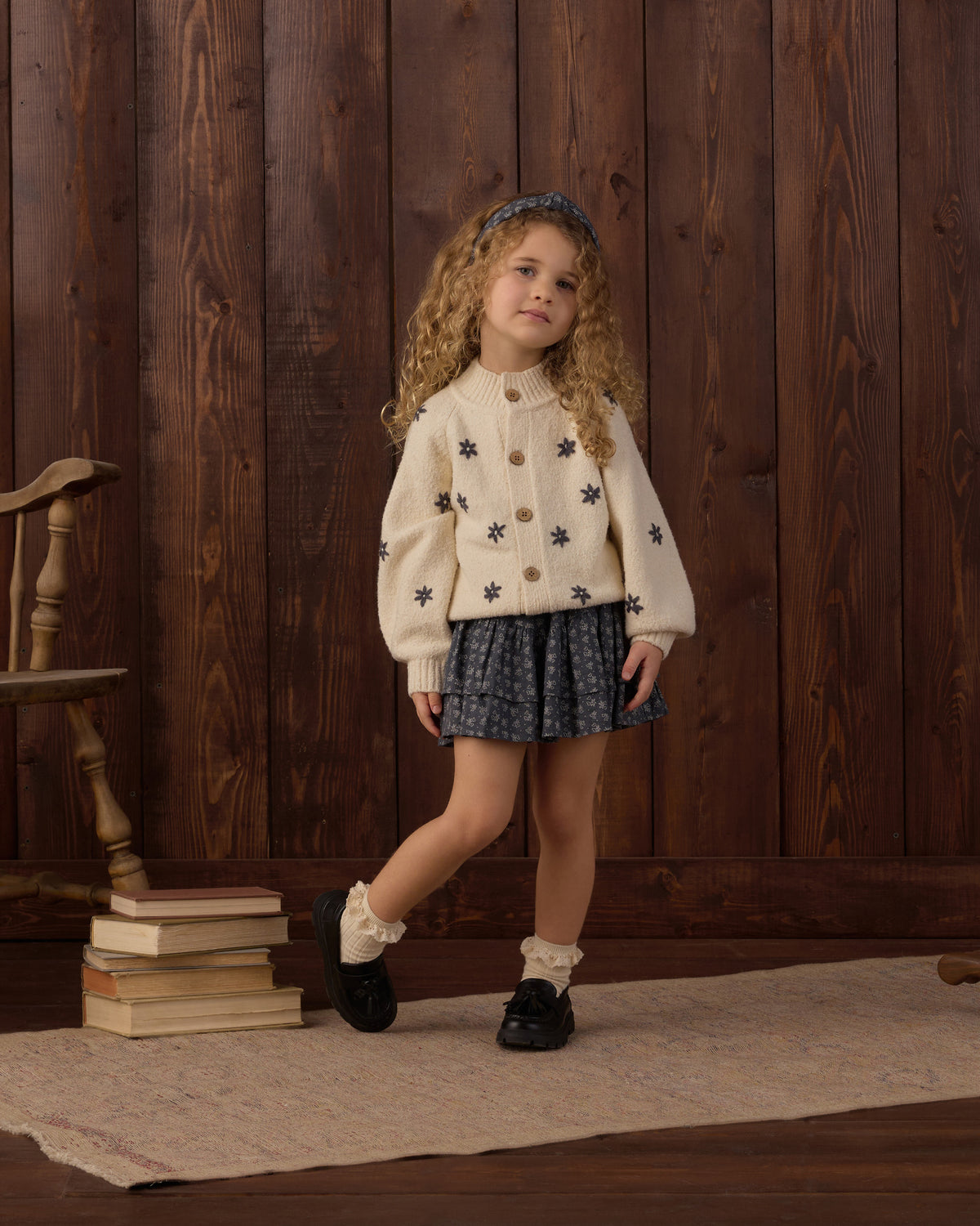 A young girl with curly light brown hair stands in a rustic room with dark wood paneling, wearing a cream knit cardigan with embroidered dark blue star motifs, a dark blue patterned pleated skirt, white ruffled socks, and black shoes.