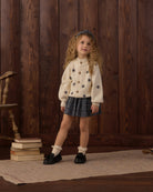 A young girl with curly hair stands in front of a dark wood plank wall, wearing a cream knit cardigan with black star motifs, a dark patterned pleated skirt, white ruffled socks, and black loafers.