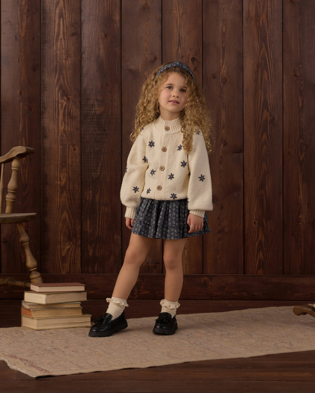 A young girl with curly hair stands in front of a dark wood plank wall, wearing a cream knit cardigan with black star motifs, a dark patterned pleated skirt, white ruffled socks, and black loafers.