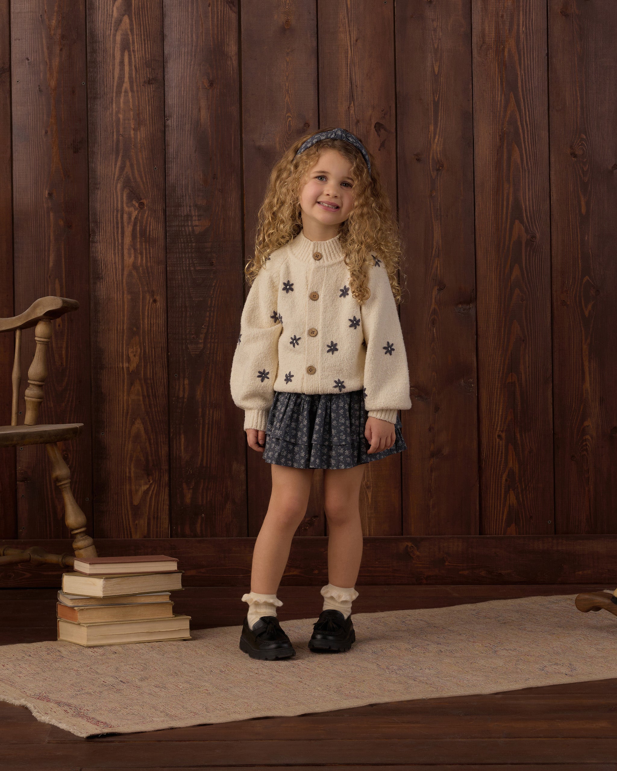 A young girl with curly hair stands in front of a dark wood plank wall wearing a cream knit cardigan with black star patterns, a dark patterned pleated skirt, white socks, and black shoes.