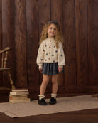A young girl with curly hair stands in front of a dark wood plank wall wearing a cream knit cardigan with black star patterns, a dark patterned pleated skirt, white socks, and black shoes.