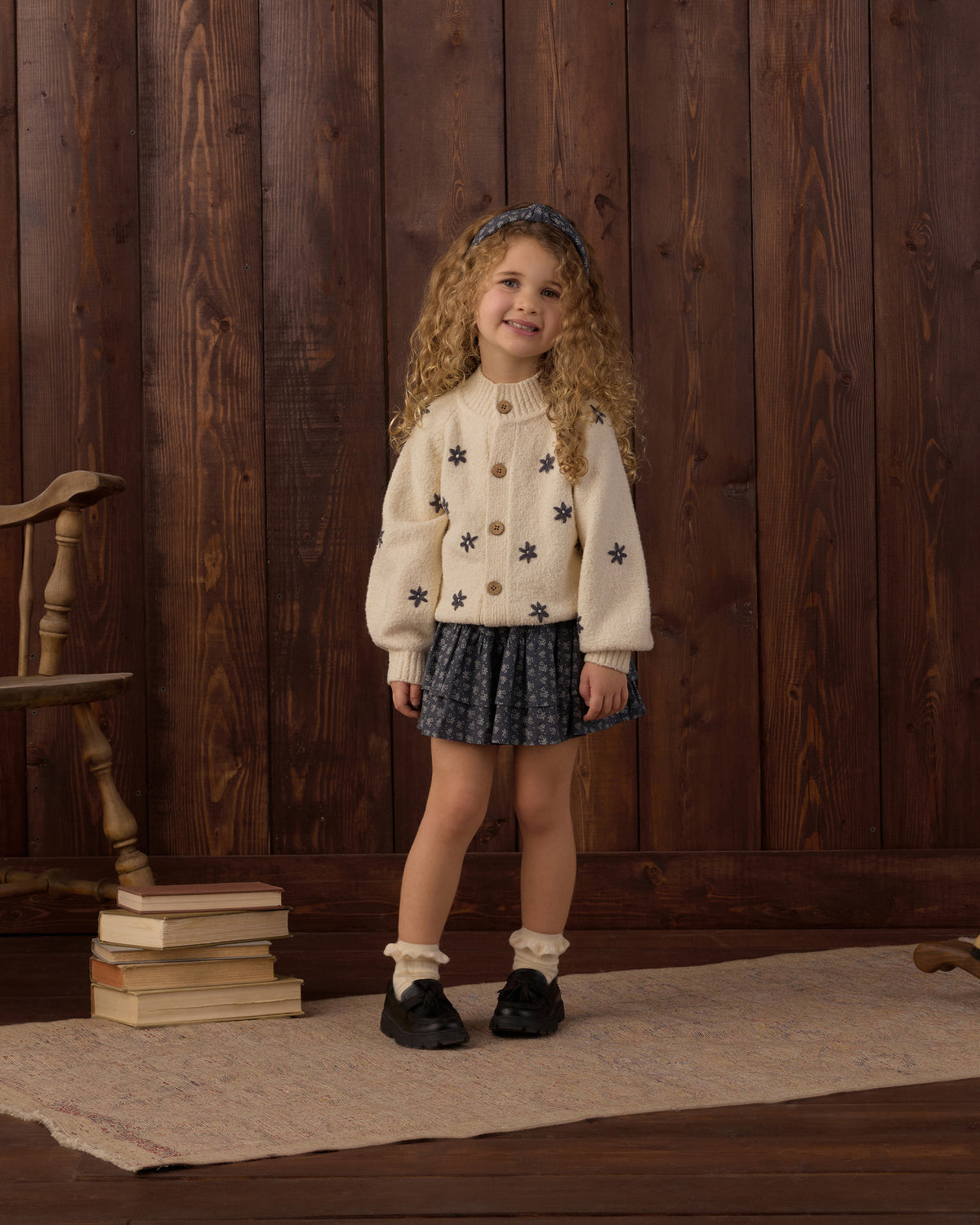 A young girl with curly hair stands in front of a dark wood plank wall wearing a cream knit cardigan with black star patterns, a dark patterned pleated skirt, white socks, and black shoes.