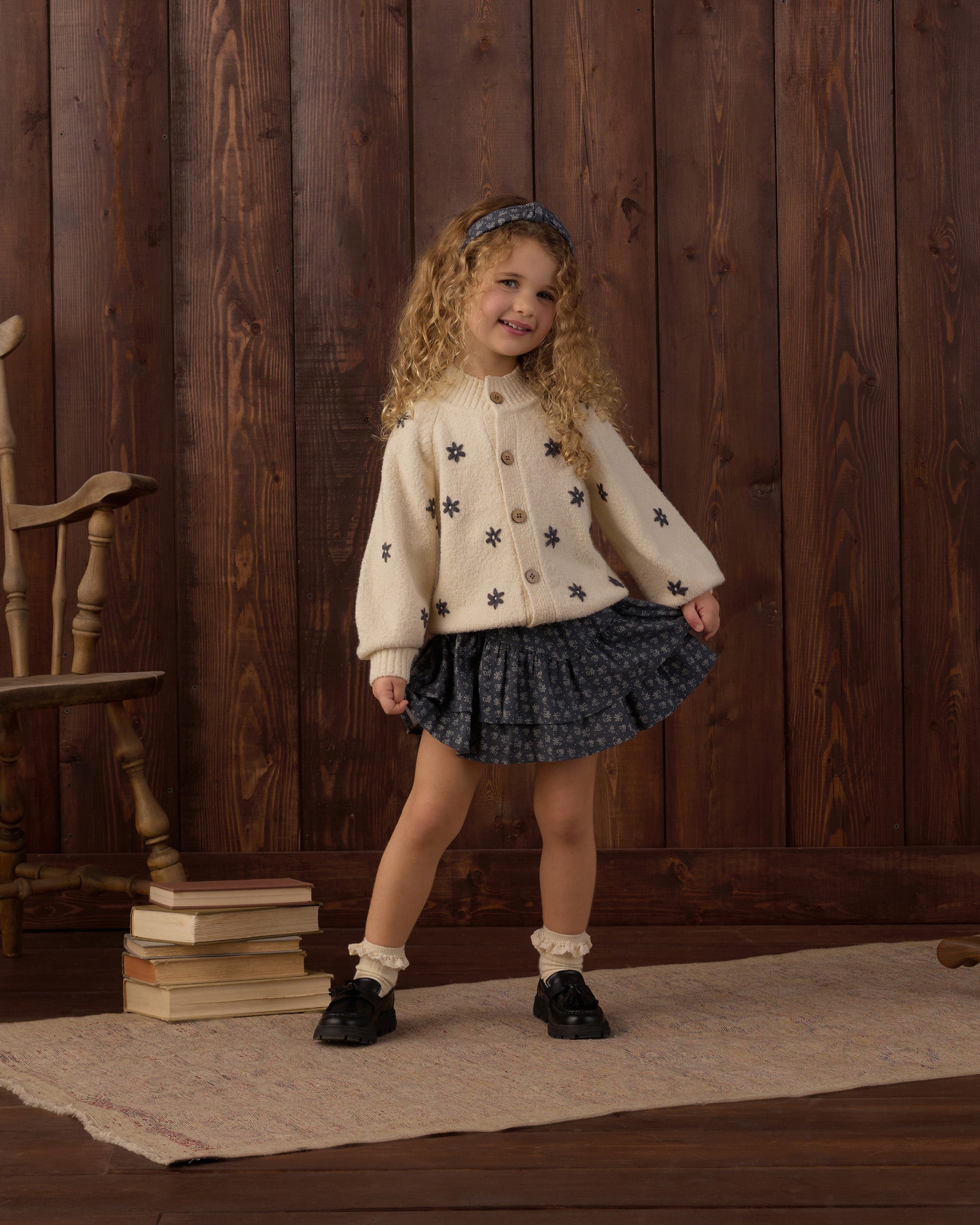 A young girl with curly blonde hair stands in a rustic room with dark wood paneling, wearing a cream cardigan with black star patterns, a dark blue patterned pleated skirt, white socks, and black shoes.