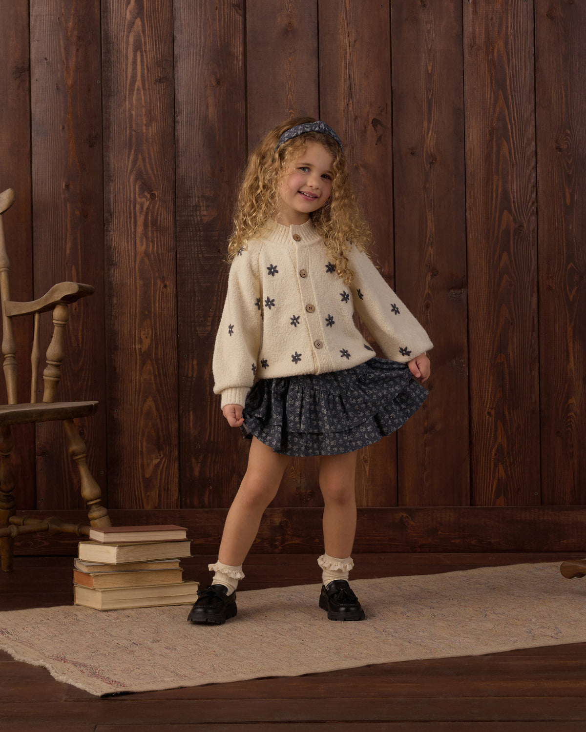 A young girl with curly blonde hair stands in a rustic room with dark wood paneling, wearing a cream cardigan with black star patterns, a dark blue patterned pleated skirt, white socks, and black shoes.