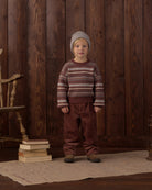 A young child stands against a dark wood plank wall wearing a knit beanie, a long-sleeve crewneck sweater with horizontal stripes in brown, beige, and white, and brown pants.