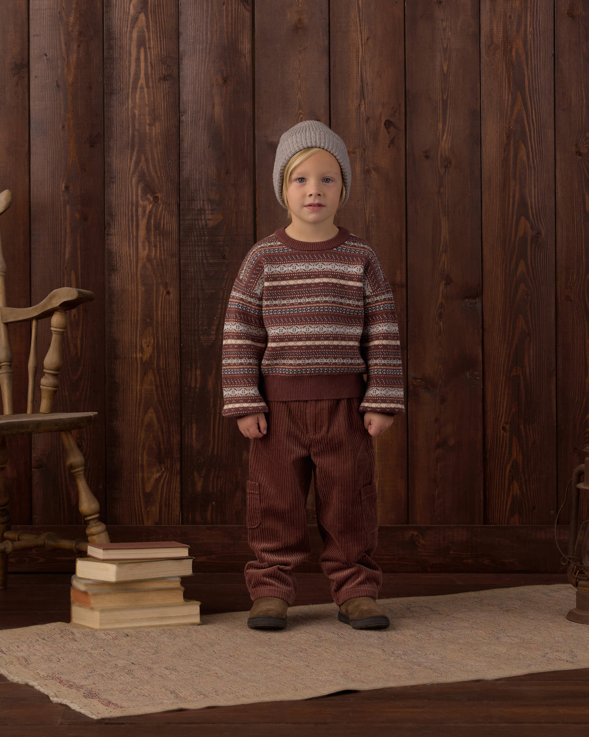 A young child stands against a dark wood plank wall wearing a knit beanie, a long-sleeve crewneck sweater with horizontal stripes in brown, beige, and white, and brown pants.
