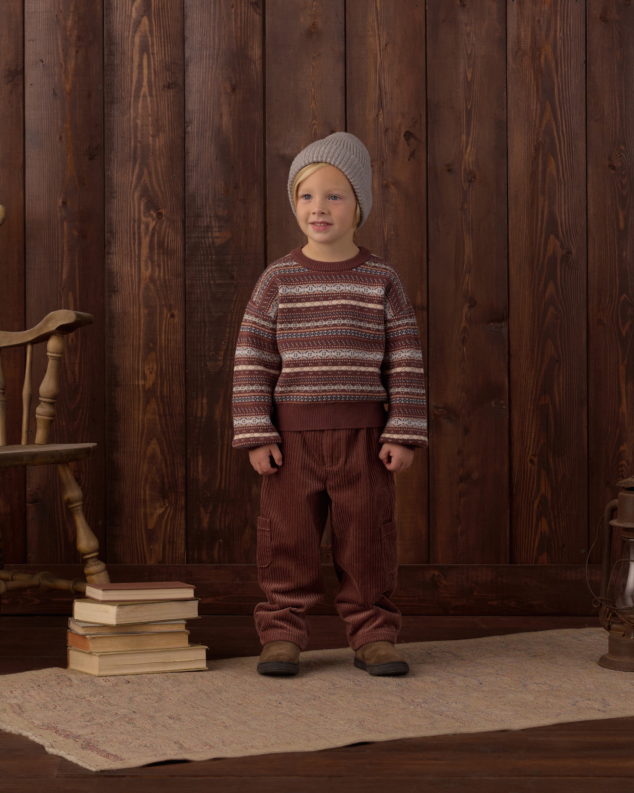 A young child stands in front of a dark wood plank wall wearing a knit sweater with horizontal brown, beige, and burgundy stripes, a gray knit beanie, and brown pants. A stack of books and a wooden chair are visible on the left, and a rug covers the floor.