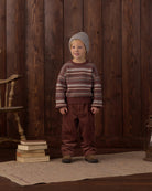 A young child stands in front of a dark wood plank wall wearing a knit sweater with horizontal brown, beige, and burgundy stripes, a gray knit beanie, and brown pants. A stack of books and a wooden chair are visible on the left, and a rug covers the floor.