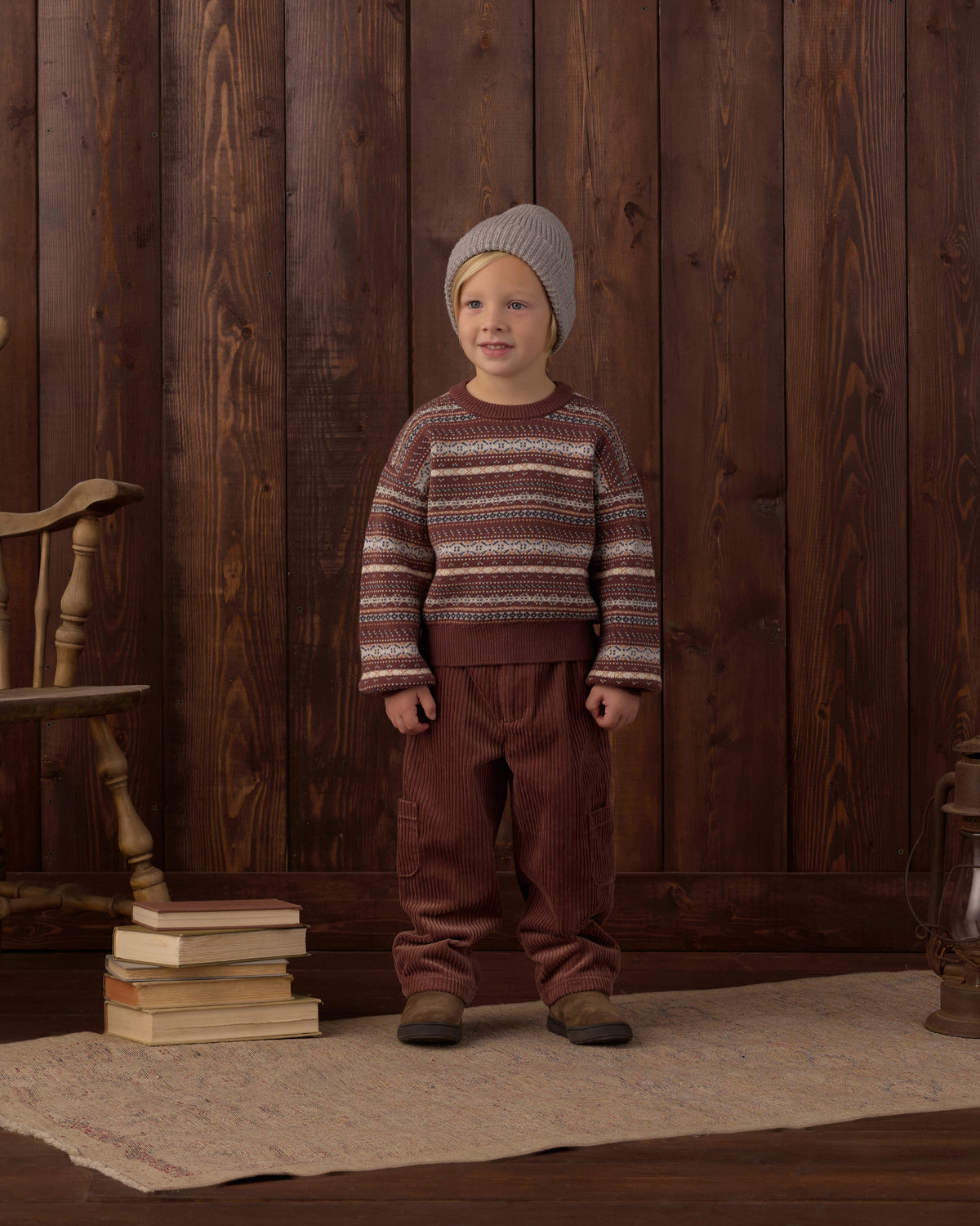 A young child stands in front of a dark wood plank wall wearing a knit sweater with horizontal brown, beige, and burgundy stripes, a gray knit beanie, and brown pants. A stack of books and a wooden chair are visible on the left, and a rug covers the floor.
