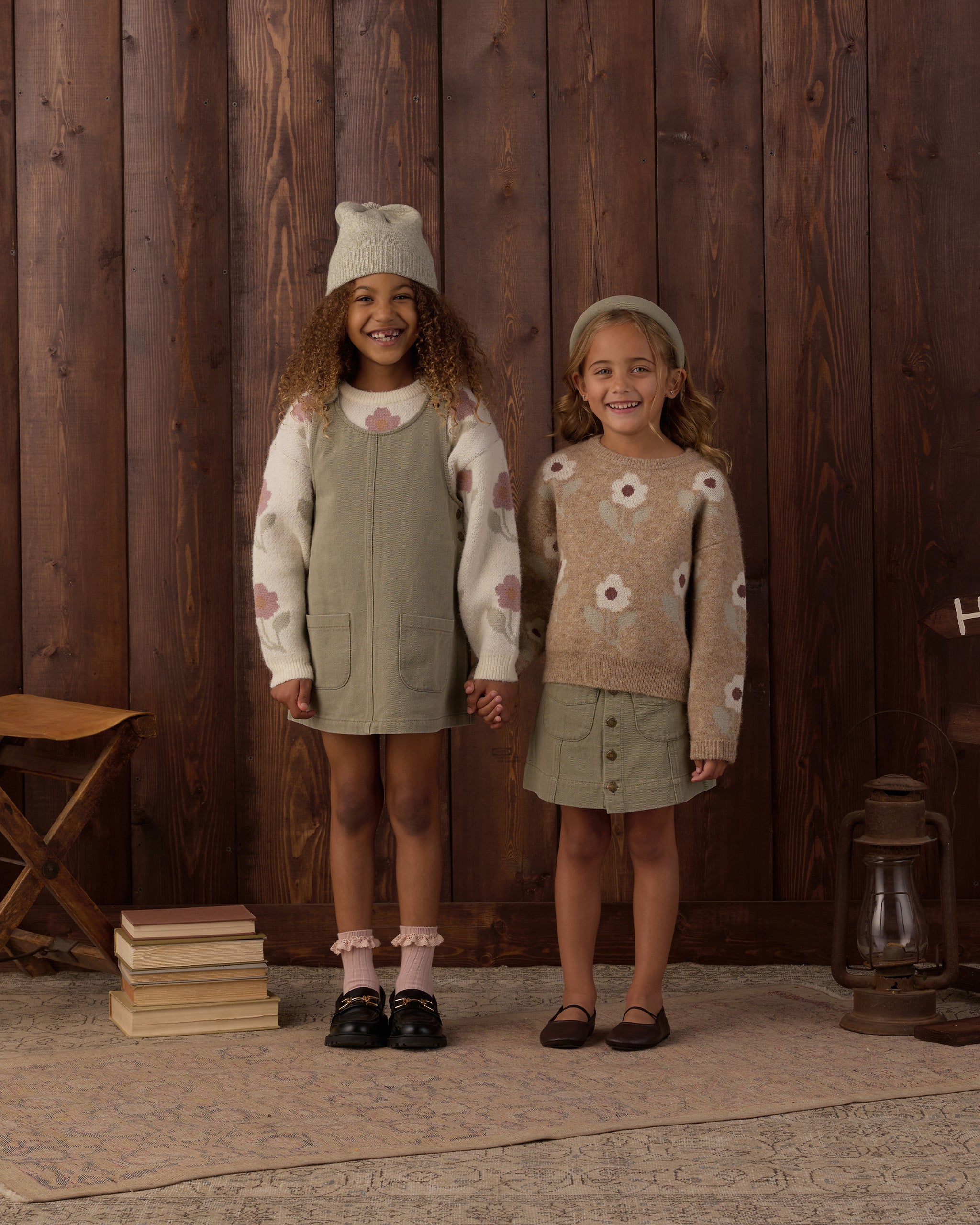 Two young girls modeling coordinated knit outfits in a rustic indoor setting with a dark wood plank wall. The girl on the left wears a cream knit sweater with pink floral motifs and a sage green pinafore dress, along with a matching cream knit hat.
