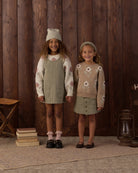 Two young girls modeling coordinated knit outfits in a rustic indoor setting with a dark wood plank wall. The girl on the left wears a cream knit sweater with pink floral motifs and a sage green pinafore dress, along with a matching cream knit hat.