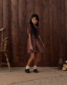 A young girl stands in front of a rustic dark wood plank wall, wearing a short-sleeve burgundy dress with a small floral pattern, white ruffled socks, and black shoes.