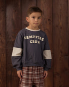 A young boy wearing a navy blue crewneck sweatshirt with 'CAMPFIRE CREW' printed in beige on the chest, featuring beige color-blocked sleeves, paired with brown and red plaid pants, standing against a dark wood plank background.