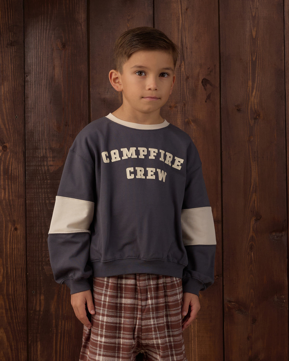 A young boy wearing a navy blue crewneck sweatshirt with 'CAMPFIRE CREW' printed in beige on the chest, featuring beige color-blocked sleeves, paired with brown and red plaid pants, standing against a dark wood plank background.