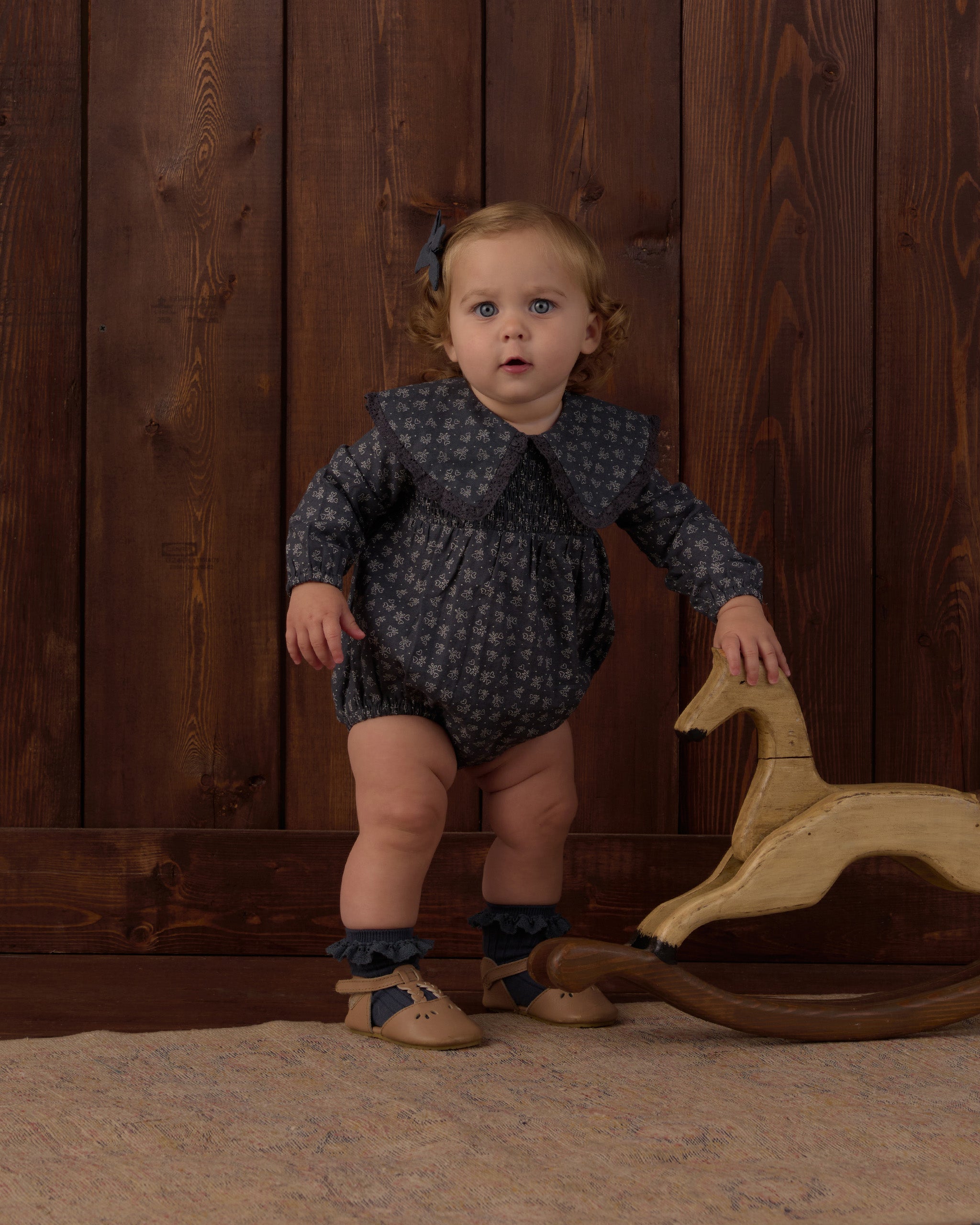 A toddler stands indoors against a dark wood plank wall, wearing a dark navy long-sleeve romper with a small light pattern, ruffled collar, ruffled socks, and tan shoes. A wooden rocking horse is beside the child.