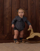 A toddler stands indoors against a dark wood plank wall, wearing a dark navy long-sleeve romper with a small light pattern, ruffled collar, ruffled socks, and tan shoes. A wooden rocking horse is beside the child.