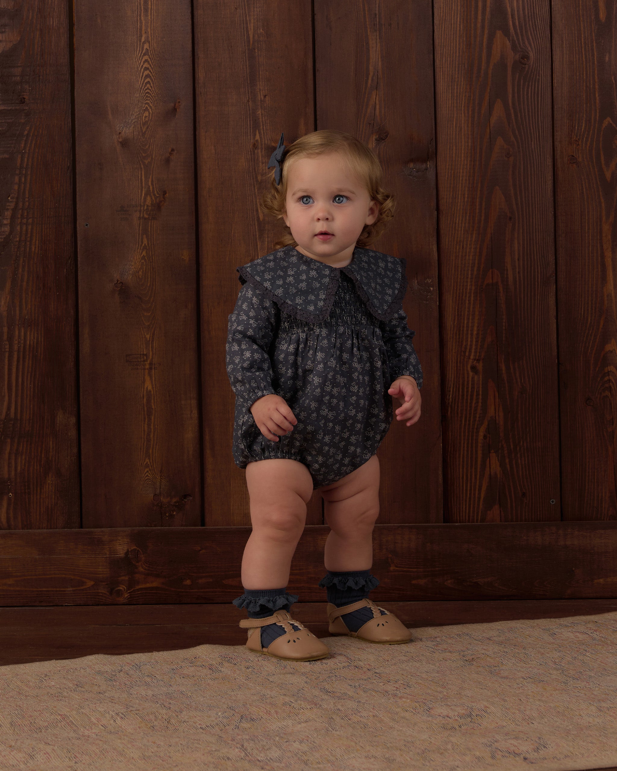 A toddler stands against a dark wood plank wall wearing a dark navy long-sleeve romper with a small light pattern, ruffled collar, ruffled leg cuffs, tan shoes, and a small blue hair bow.