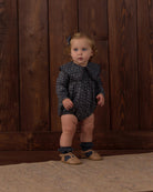A toddler stands against a dark wood plank wall wearing a dark navy long-sleeve romper with a small light pattern, ruffled collar, ruffled leg cuffs, tan shoes, and a small blue hair bow.