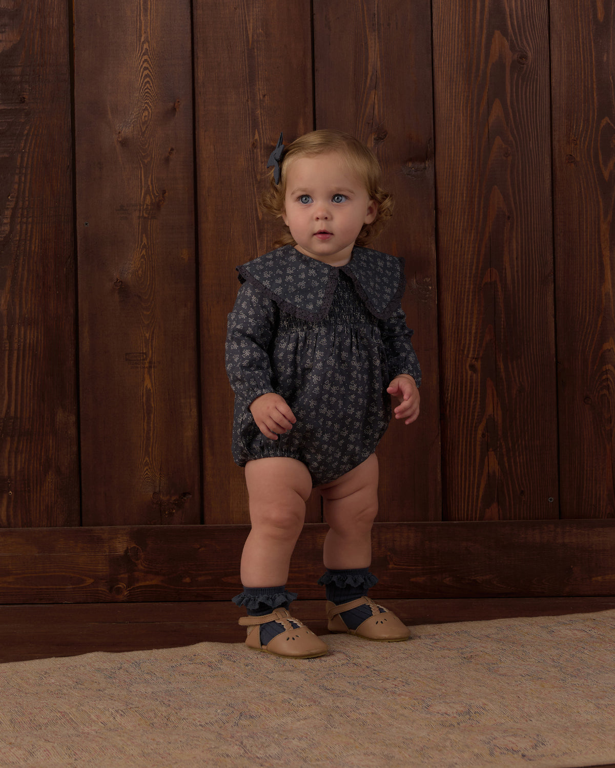 A toddler stands against a dark wood plank wall wearing a dark navy long-sleeve romper with a small light pattern, ruffled collar, ruffled leg cuffs, tan shoes, and a small blue hair bow.