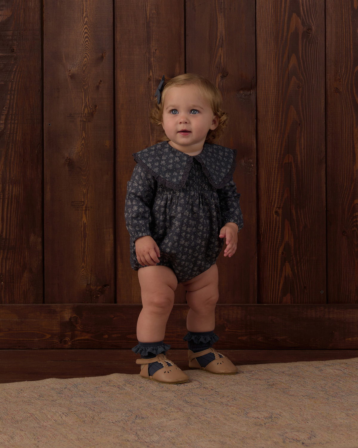 A toddler stands against a dark wood plank wall wearing a dark navy long-sleeve romper with a small light pattern, ruffled collar, ruffled leg cuffs, tan shoes, and tan socks.