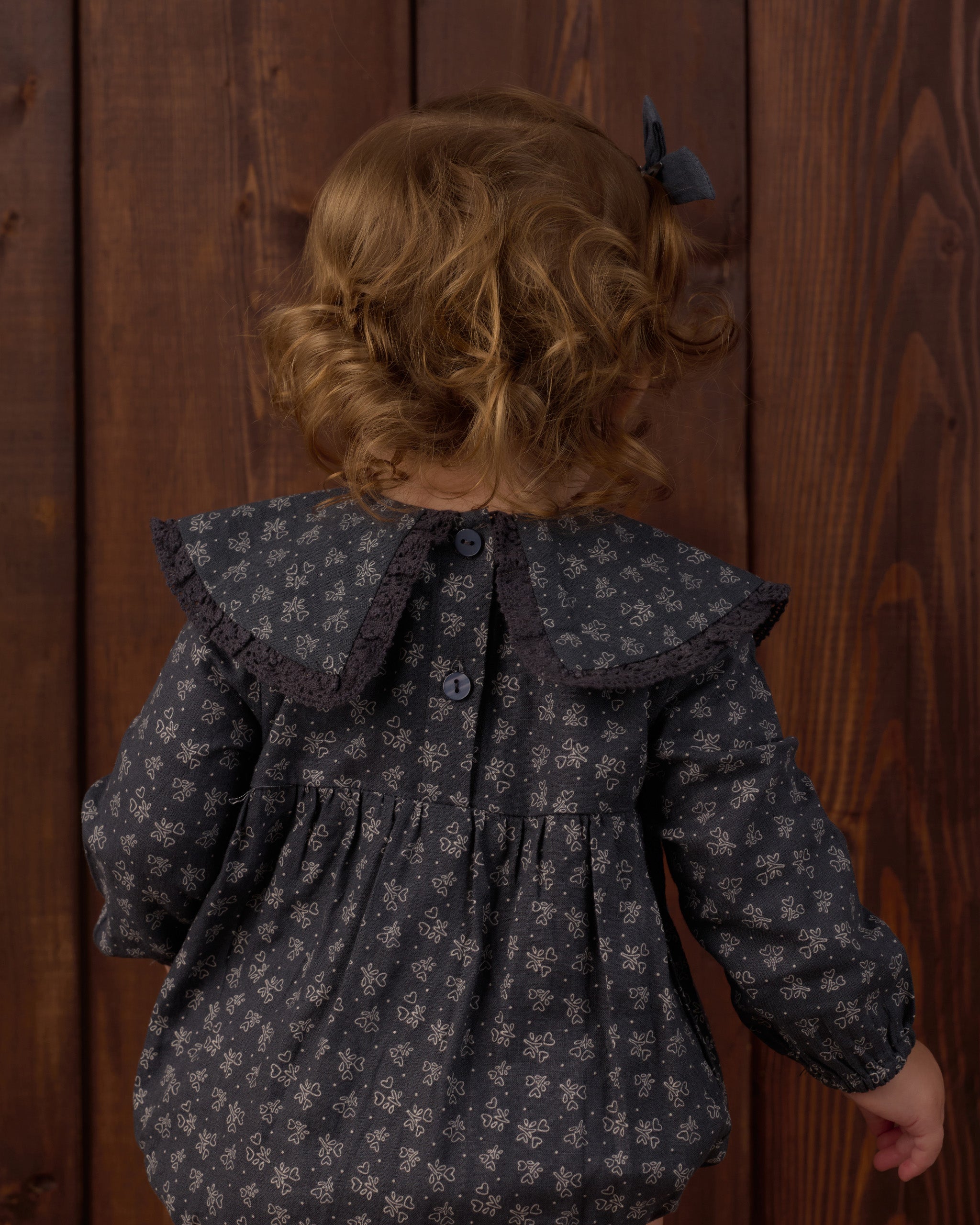 A child viewed from behind wearing a dark gray long-sleeve blouse with a small white floral pattern, a large ruffled collar, and a dark bow in their hair, standing against a dark wood plank background.
