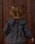 A child viewed from behind wearing a dark gray long-sleeve blouse with a small white floral pattern, a large ruffled collar, and a dark bow in their hair, standing against a dark wood plank background.