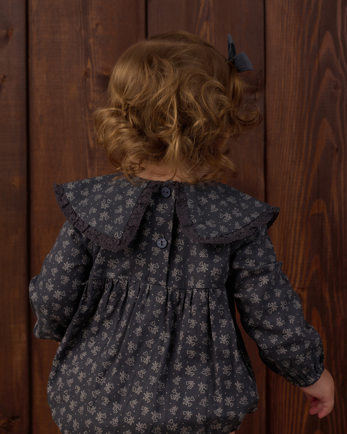 A child viewed from behind wearing a dark gray long-sleeve blouse with a small white floral pattern, a large ruffled collar, and a dark bow in their hair, standing against a dark wood plank background.
