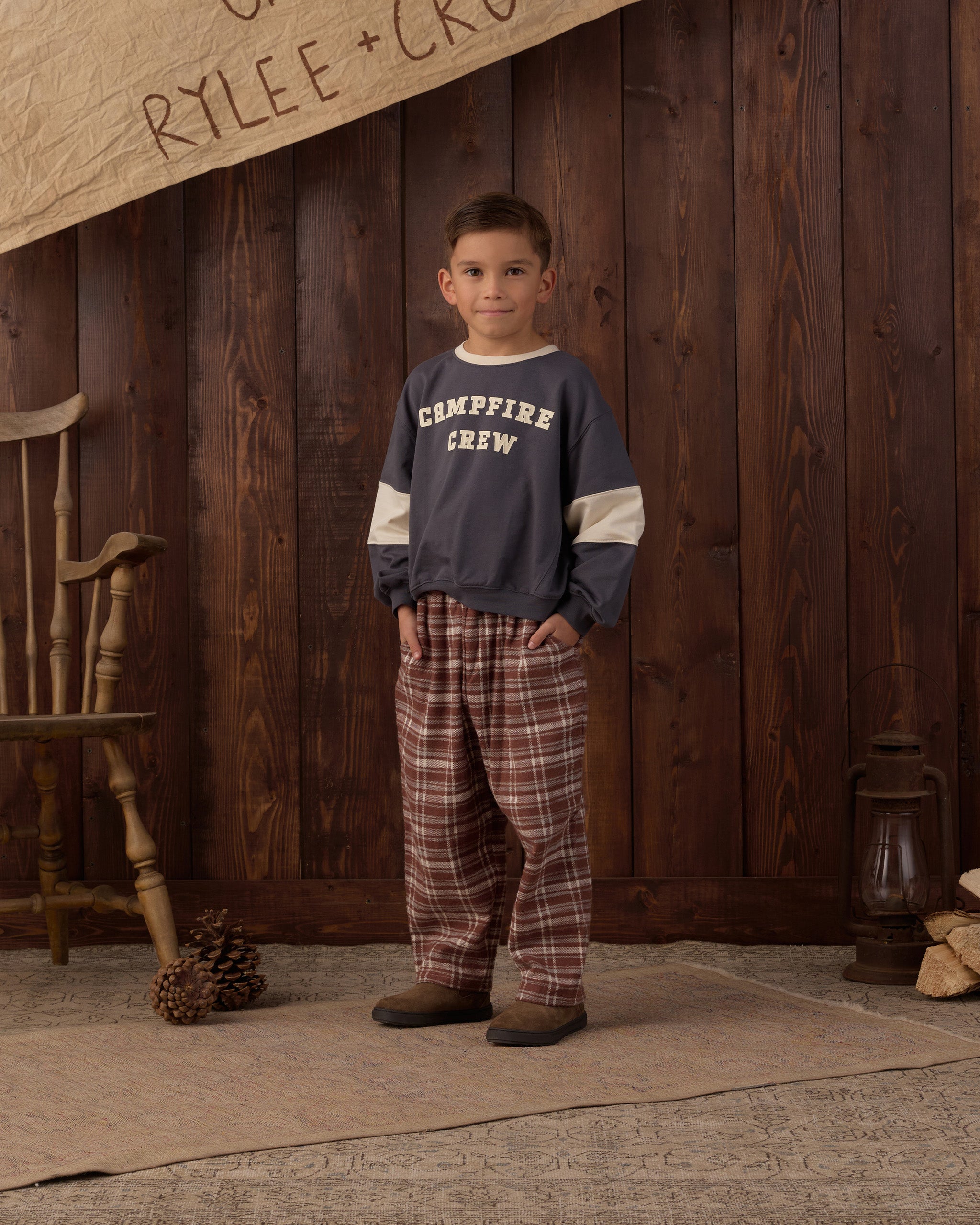 A young boy stands in front of a rustic dark wood plank wall wearing a navy blue long-sleeve sweatshirt with cream sleeves and 'CAMPFIRE CREW' printed in white on the chest, paired with brown and red plaid pajama pants.