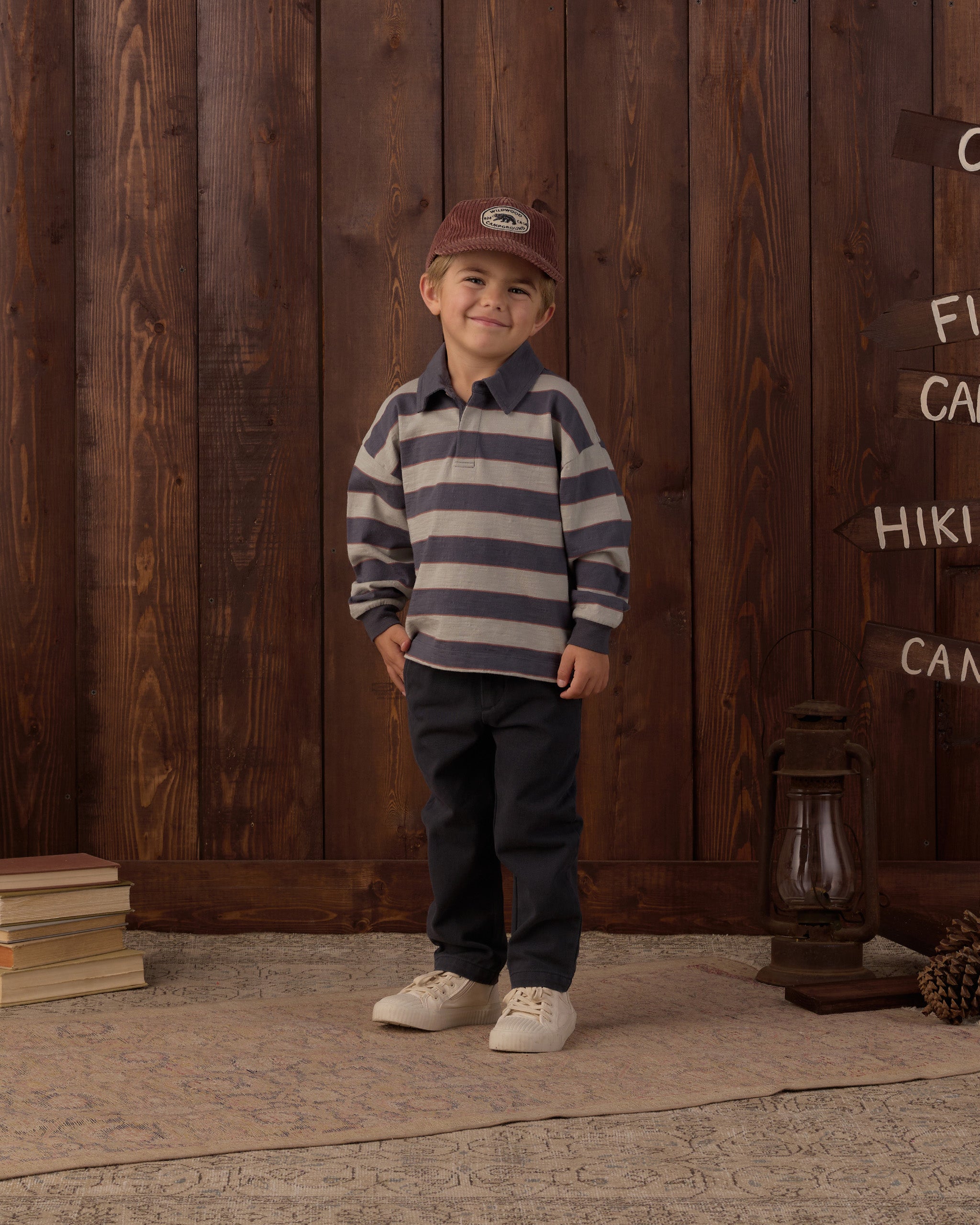 A young boy stands in front of a rustic dark wood plank wall wearing a navy and beige horizontally striped long-sleeve polo shirt, dark pants, white sneakers, and a brown baseball cap.