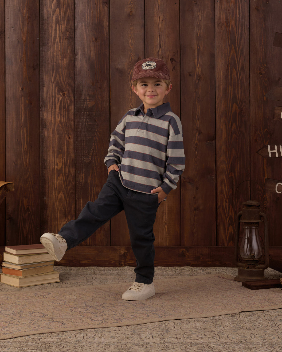 A young child wearing a navy and gray striped long-sleeve polo shirt, dark pants, white sneakers, and a brown cap, posing in front of a rustic dark wood plank wall.