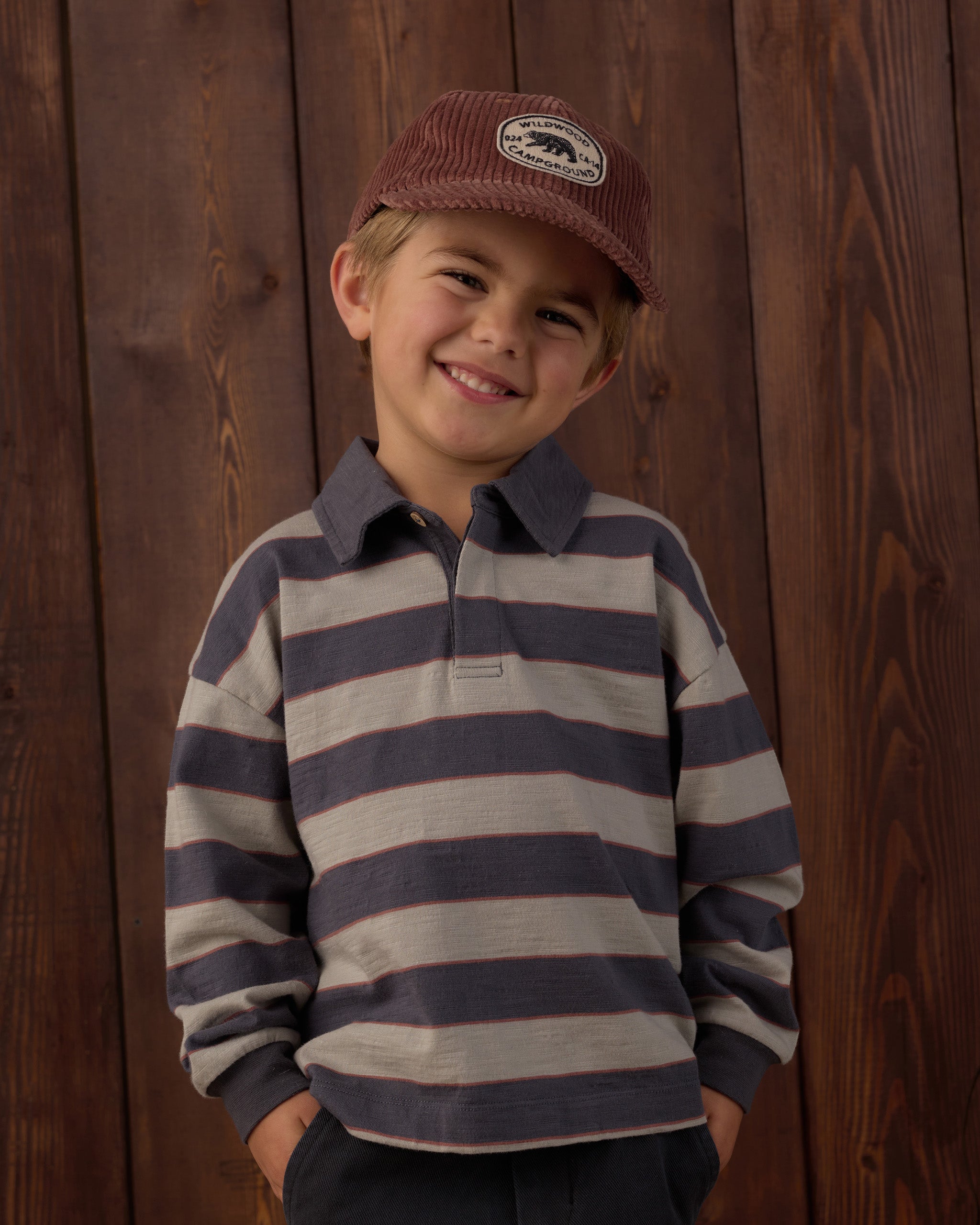 A young boy smiling, wearing a long-sleeve rugby-style shirt with horizontal navy and beige stripes, a brown corduroy cap, and dark pants, standing against a dark wood plank wall.