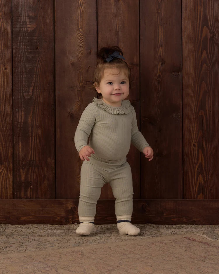 A toddler wearing a neutral sage-green ribbed long-sleeve top and matching leggings with a ruffled collar detail.