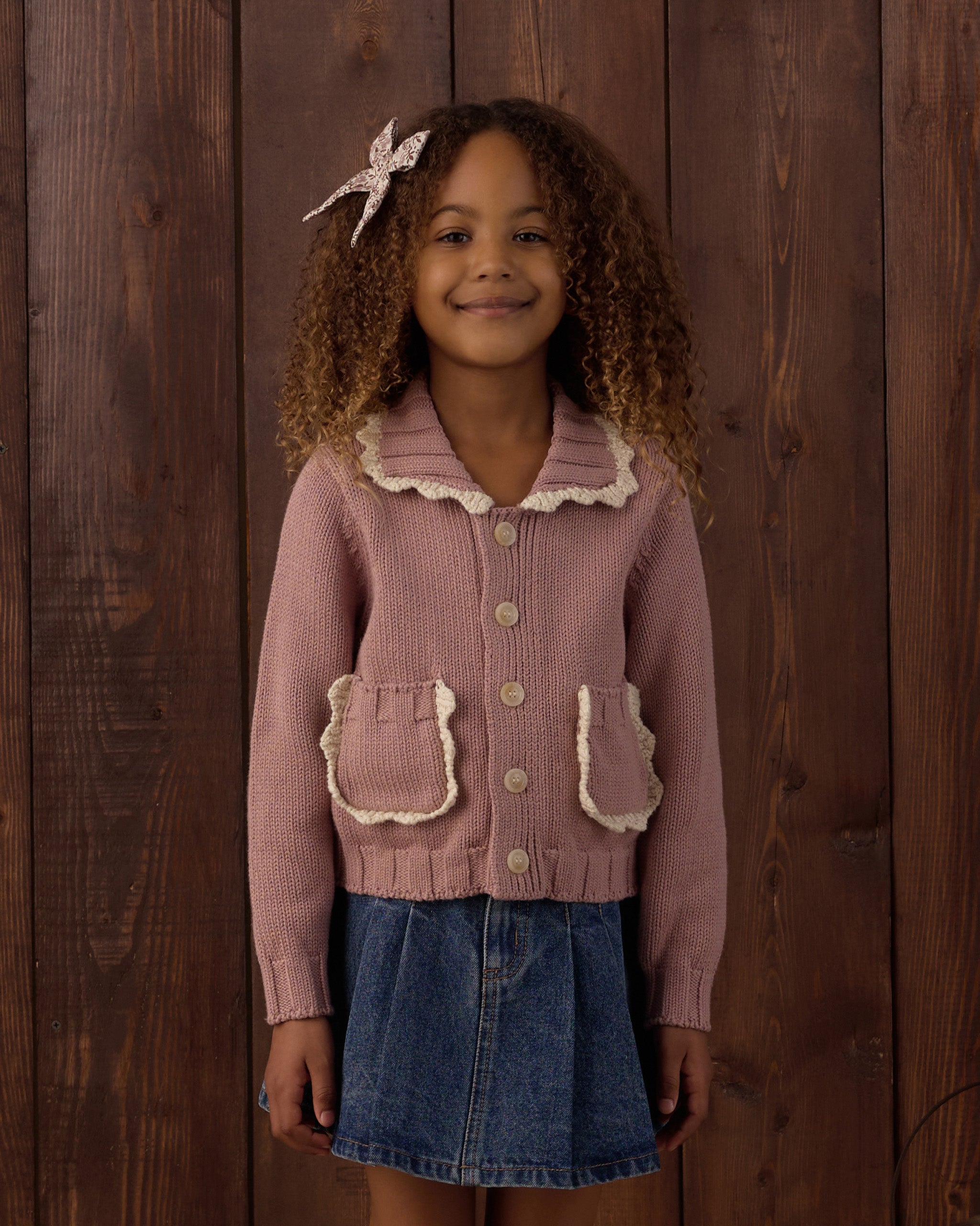 A young girl with curly hair wearing a dusty pink ribbed knit cardigan with cream ruffle trim and a denim skirt, standing against a dark wood plank background.
