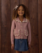 A young girl with curly hair wearing a dusty pink ribbed knit cardigan with cream ruffle trim and a denim skirt, standing against a dark wood plank background.