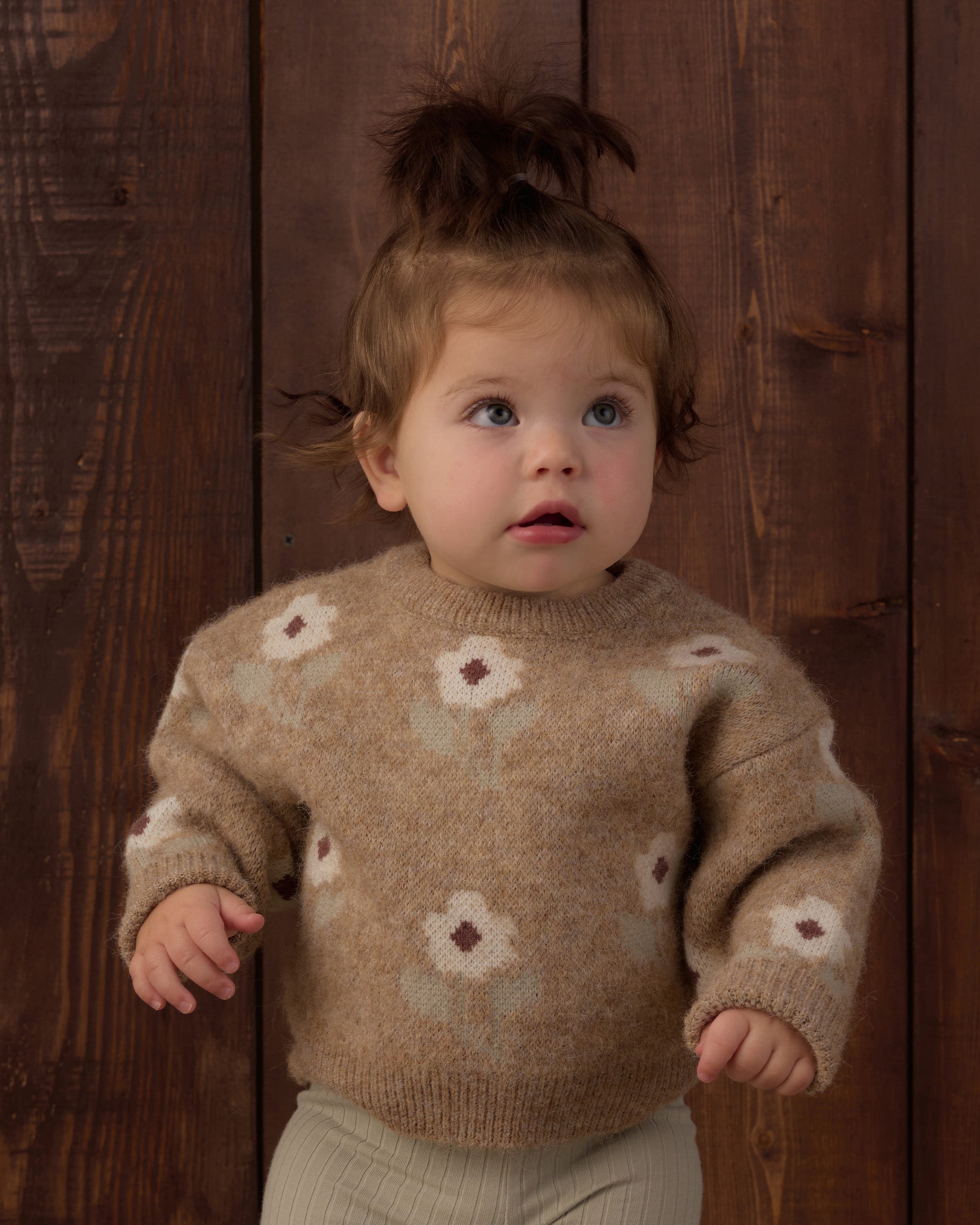 A toddler wearing a beige knit sweater with a floral pattern, standing against a dark wood plank background.