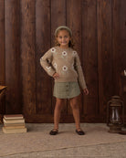 A young girl stands in front of a dark wood plank wall wearing a fuzzy beige sweater with white flower motifs, a sage green mini skirt, a matching green beanie, and black flats. A stack of books sits on the left, and a lantern rests on the right.