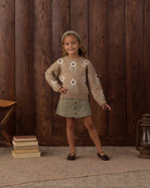 A young girl stands in front of a dark wood plank wall wearing a beige knit sweater with white and brown floral motifs, a sage green skirt, and dark flats.