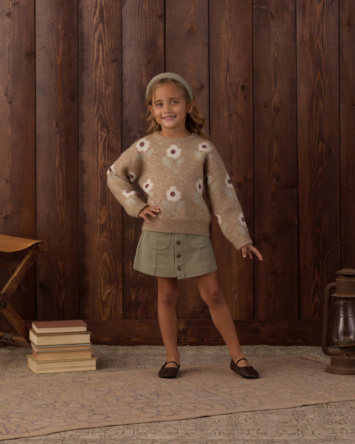A young girl stands in front of a dark wood plank wall wearing a beige knit sweater with white and brown floral motifs, a sage green skirt, and dark flats.