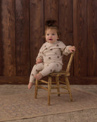A toddler girl sits on a small wooden chair in front of a dark wood plank wall, wearing a cream-colored long-sleeve pajama set with small brown floral prints. The setting includes a vintage-style wooden chair and a patterned rug.