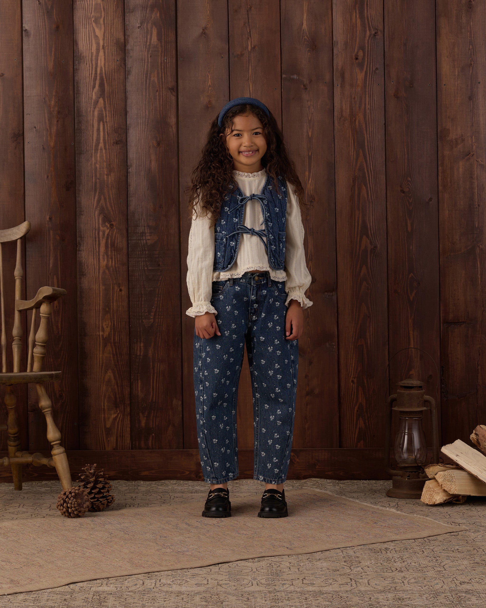 A young girl stands in front of a rustic dark wood plank wall wearing a vintage-style outfit consisting of a white long-sleeve blouse, a blue patterned vest, matching blue patterned pants, and a blue headband.