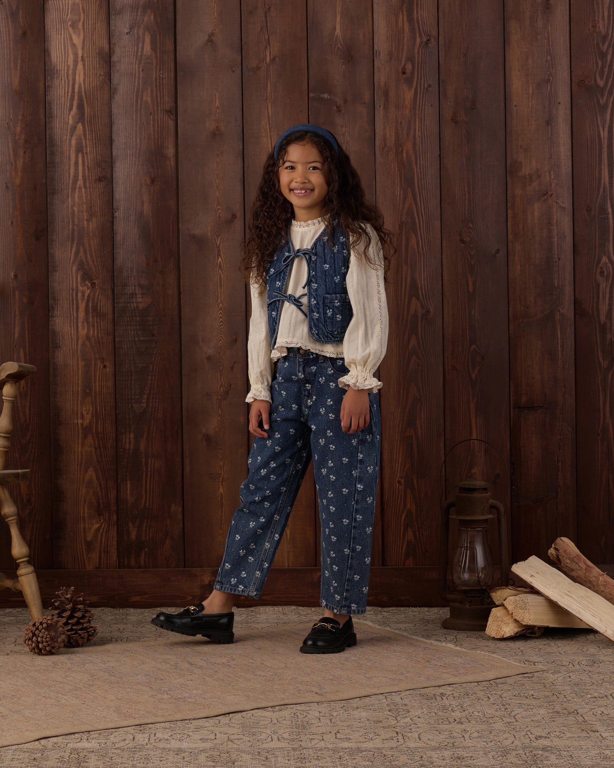 A young girl stands in front of a rustic dark wood plank wall wearing a vintage-style outfit consisting of a cream ruffled blouse, a blue floral vest, matching blue floral pants, and black shoes. The scene is styled with a wooden chair leg, pinecones, a lantern, and stacked logs on the floor.