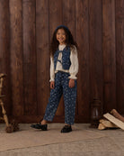 A young girl stands in front of a rustic dark wood plank wall wearing a vintage-style outfit consisting of a cream ruffled blouse, a blue floral vest, matching blue floral pants, and black shoes. The scene is styled with a wooden chair leg, pinecones, a lantern, and stacked logs on the floor.